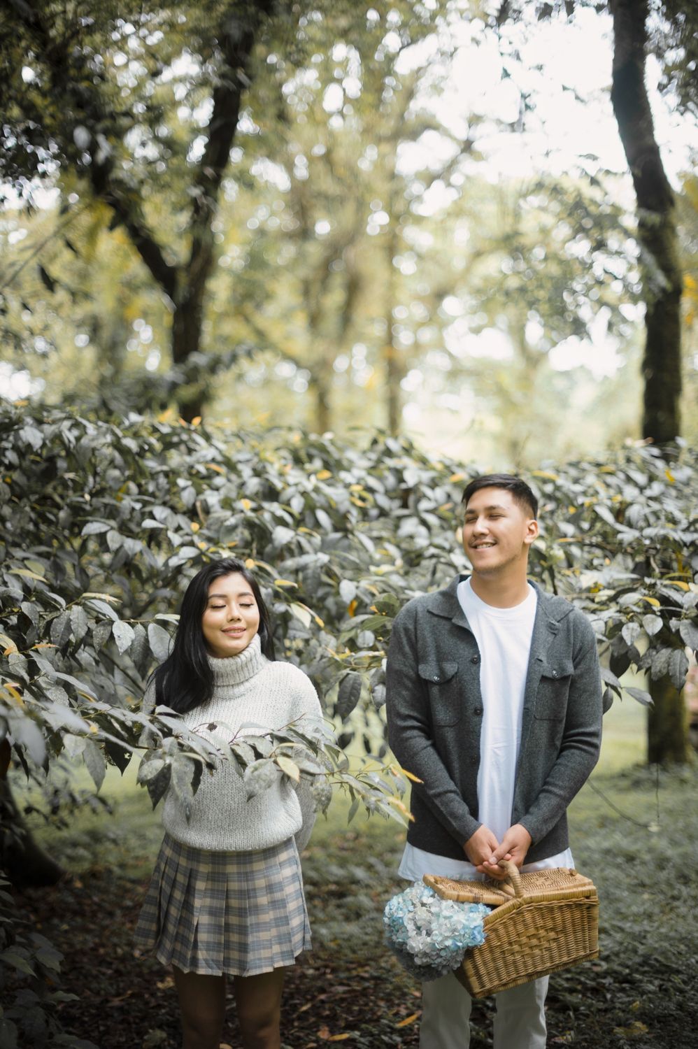 A smiling couple stands in a lush forest, woman holding branches, man carrying a wicker basket.