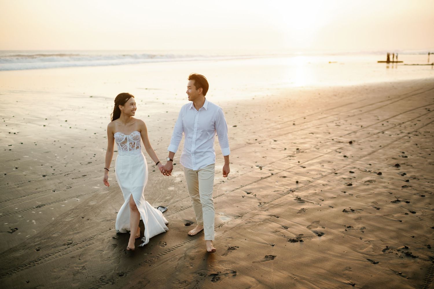 Couple in white outfits holding hands walking on a sandy beach at sunset.