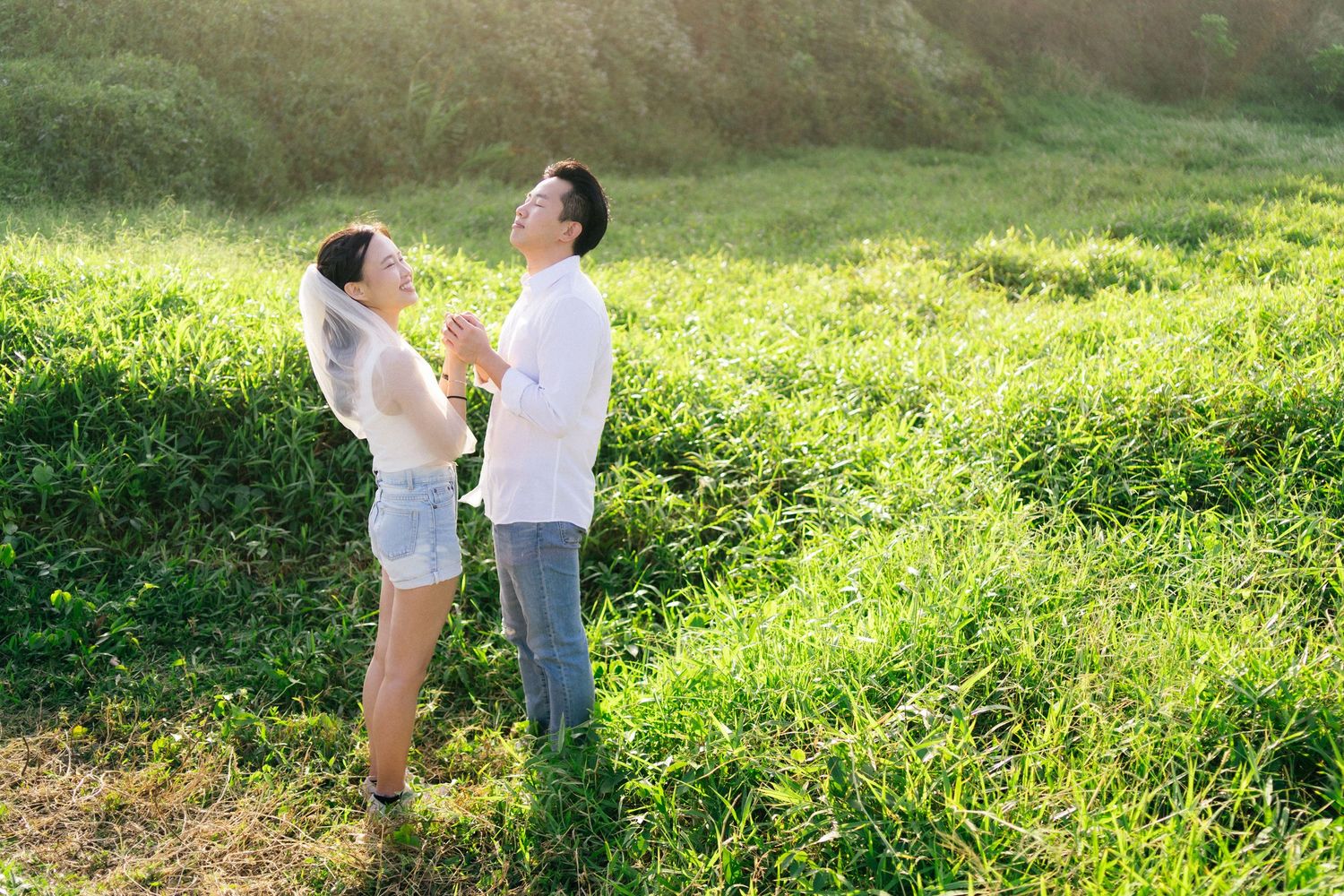 Couple holding hands and gazing at each other in a sunlit green meadow on a romantic outdoor date.
