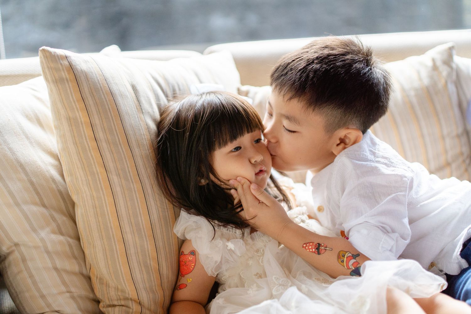 Two young Asian siblings share a sweet kiss on a cozy sofa, dressed in white outfits in a bright living room.