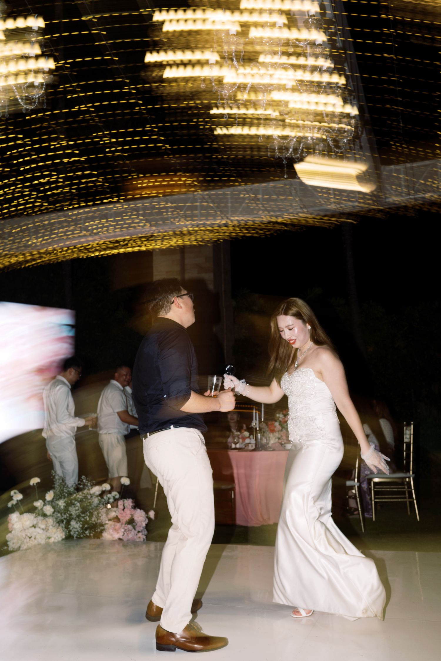 Bride and groom dancing energetically on white dance floor under glowing chandelier at elegant wedding reception.