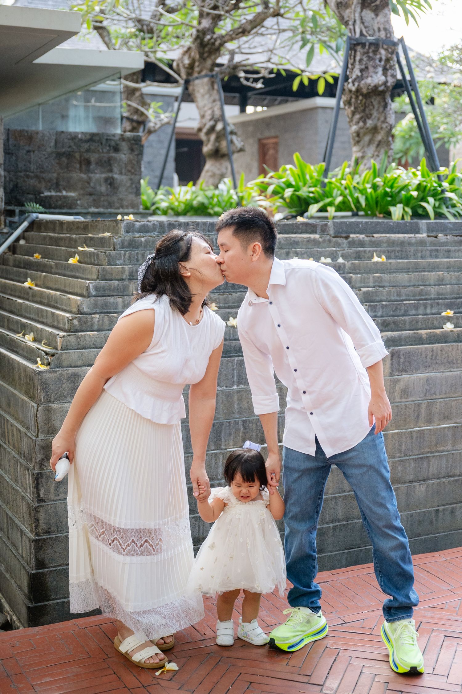 Asian couple kissing while holding their toddler daughter in white dress on outdoor steps.