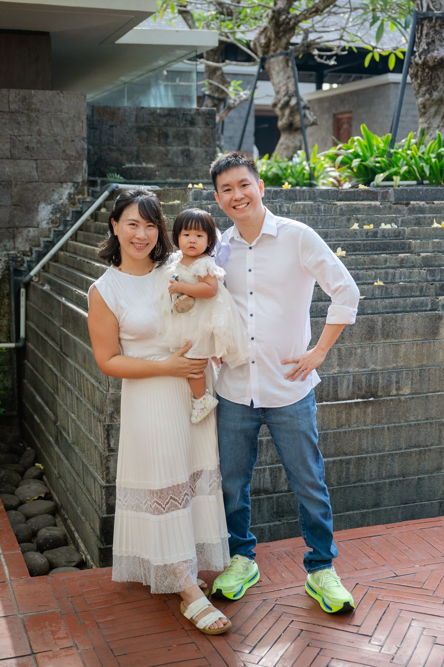 A smiling Asian family of three poses outdoors, mother in white dress holding toddler, father in white shirt and jeans.
