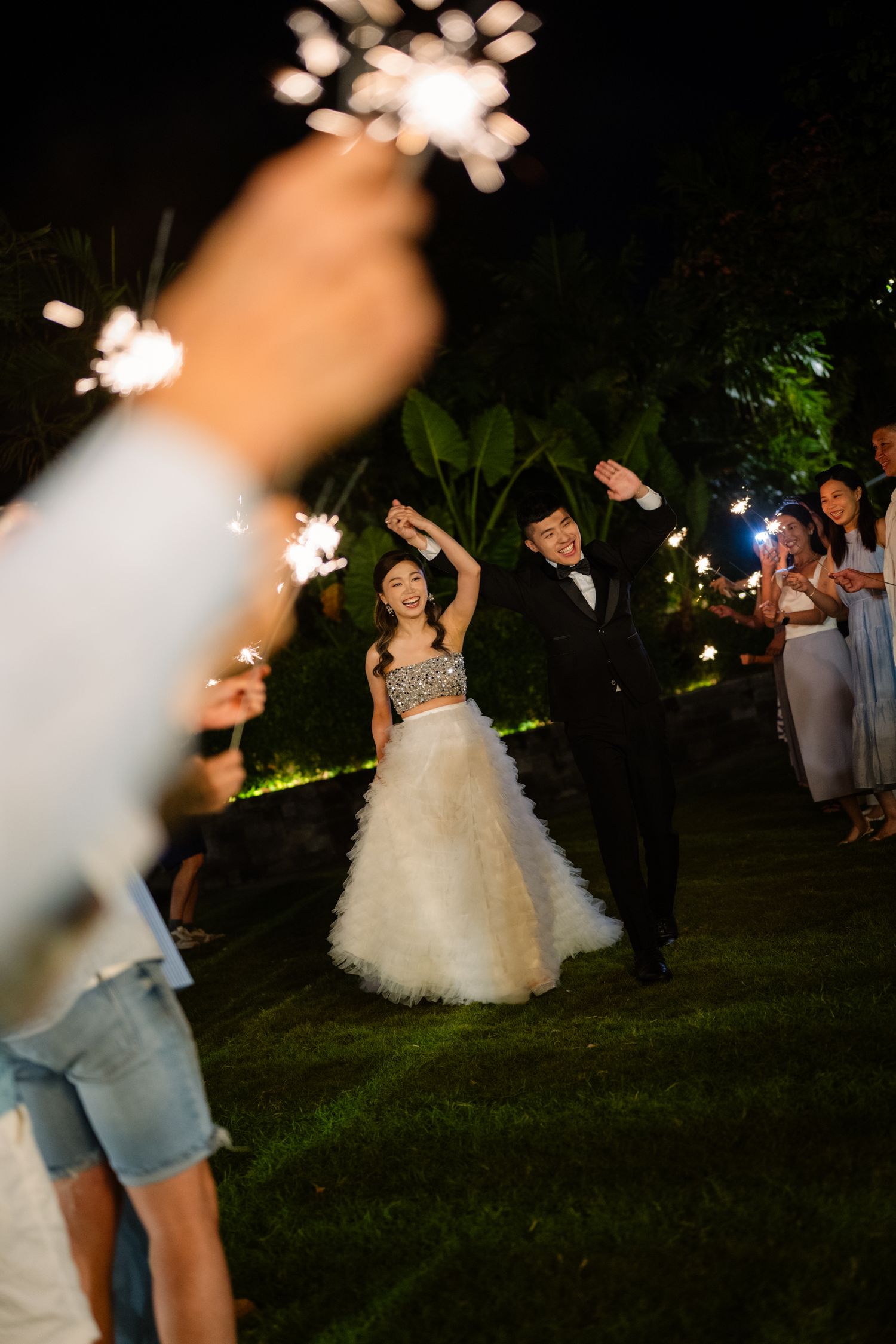 Bride in a tulle gown and groom in a tuxedo dance outdoors on a lawn at night as wedding guests hold glowing sparklers.