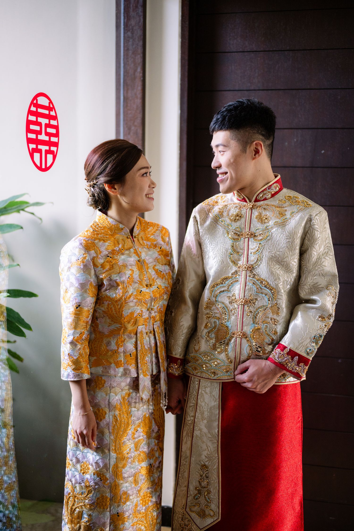 Smiling couple in traditional Chinese kua wedding attire with gold embroidery stand together beside a red double happiness symbol.