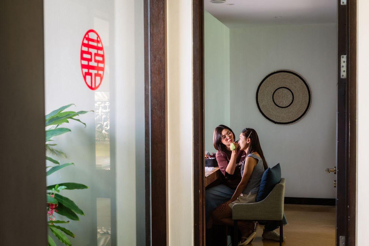 Two women sit together in a softly lit room with a red double happiness symbol on a glass door, green plants, and a round wall decoration.