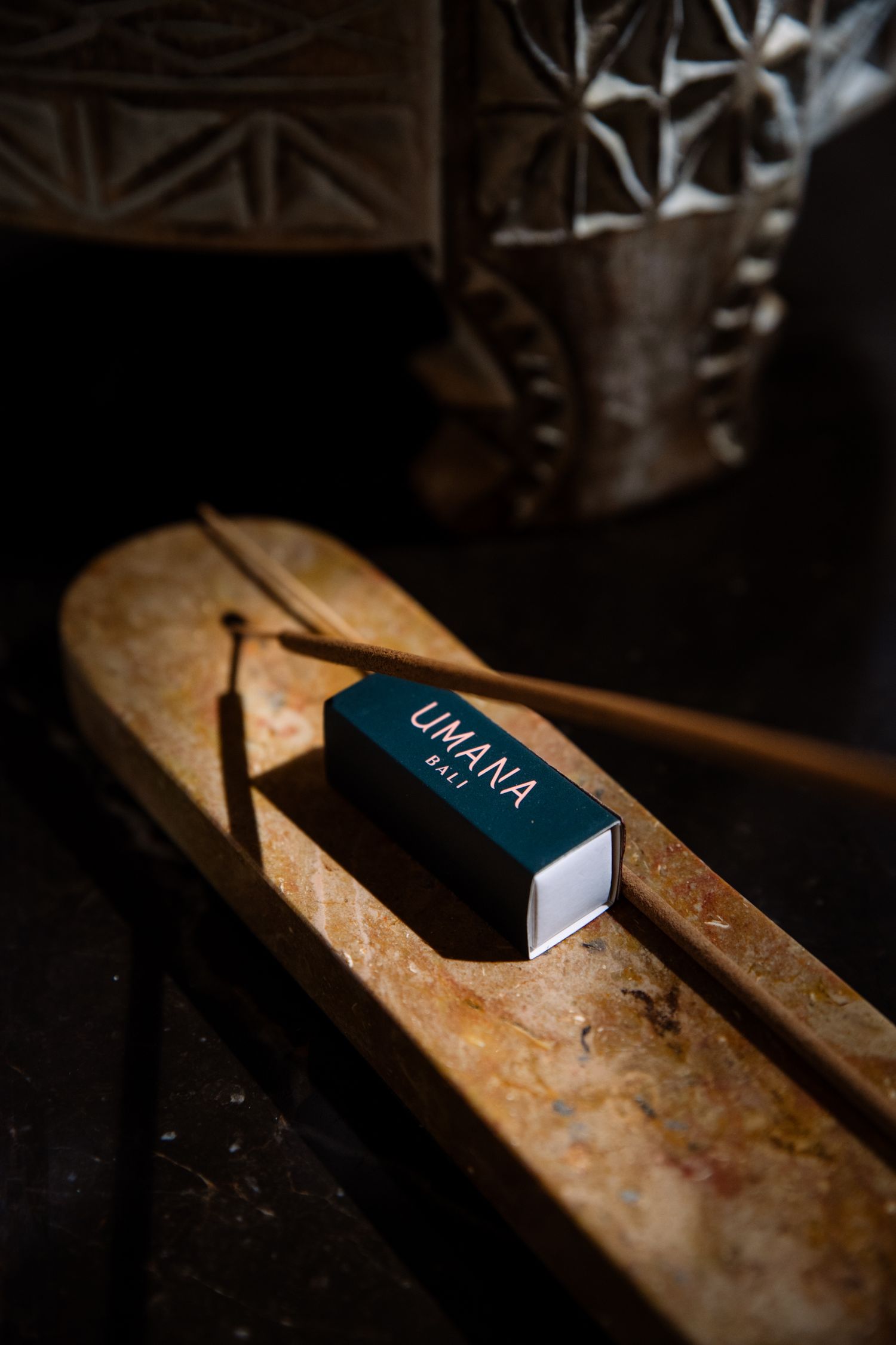 Teal Umana Bali incense box resting beside an incense stick on a polished marble holder, with carved decor softly lit in the background.