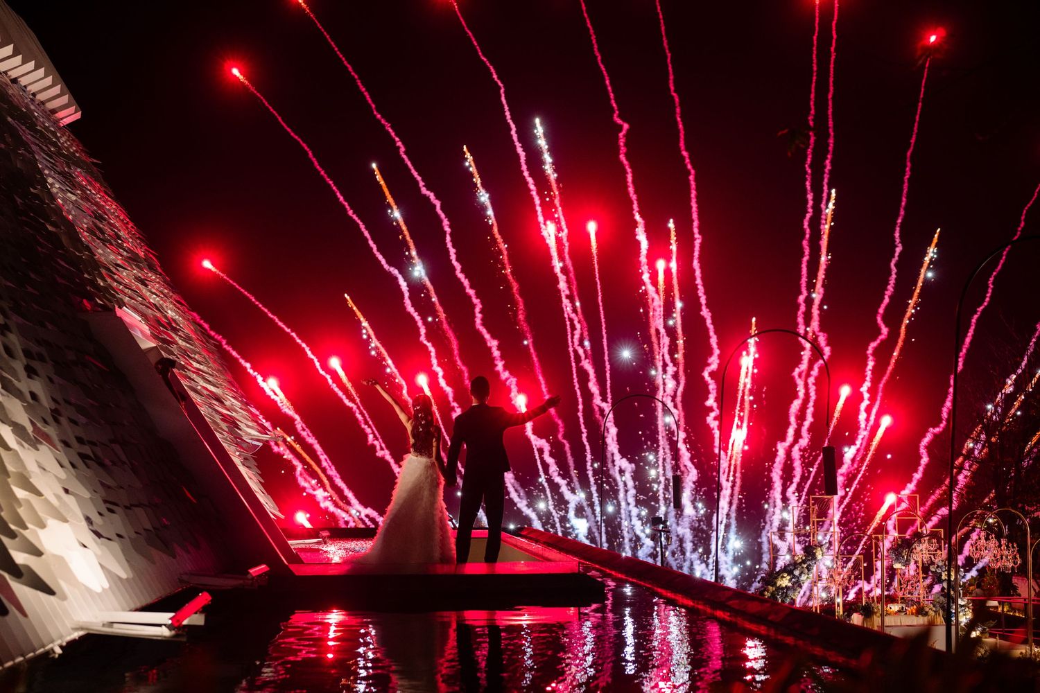 Bride and groom silhouetted on a reflective platform at night as red fireworks and sparks light up the sky during their wedding celebration.