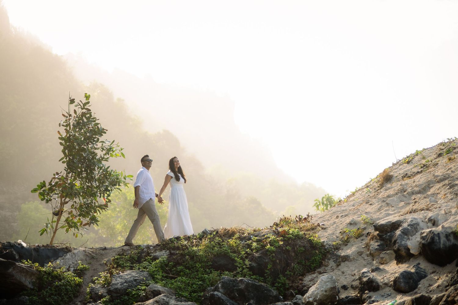 Couple holding hands walking on rocky mountain trail with misty green hills and sunlight behind.
