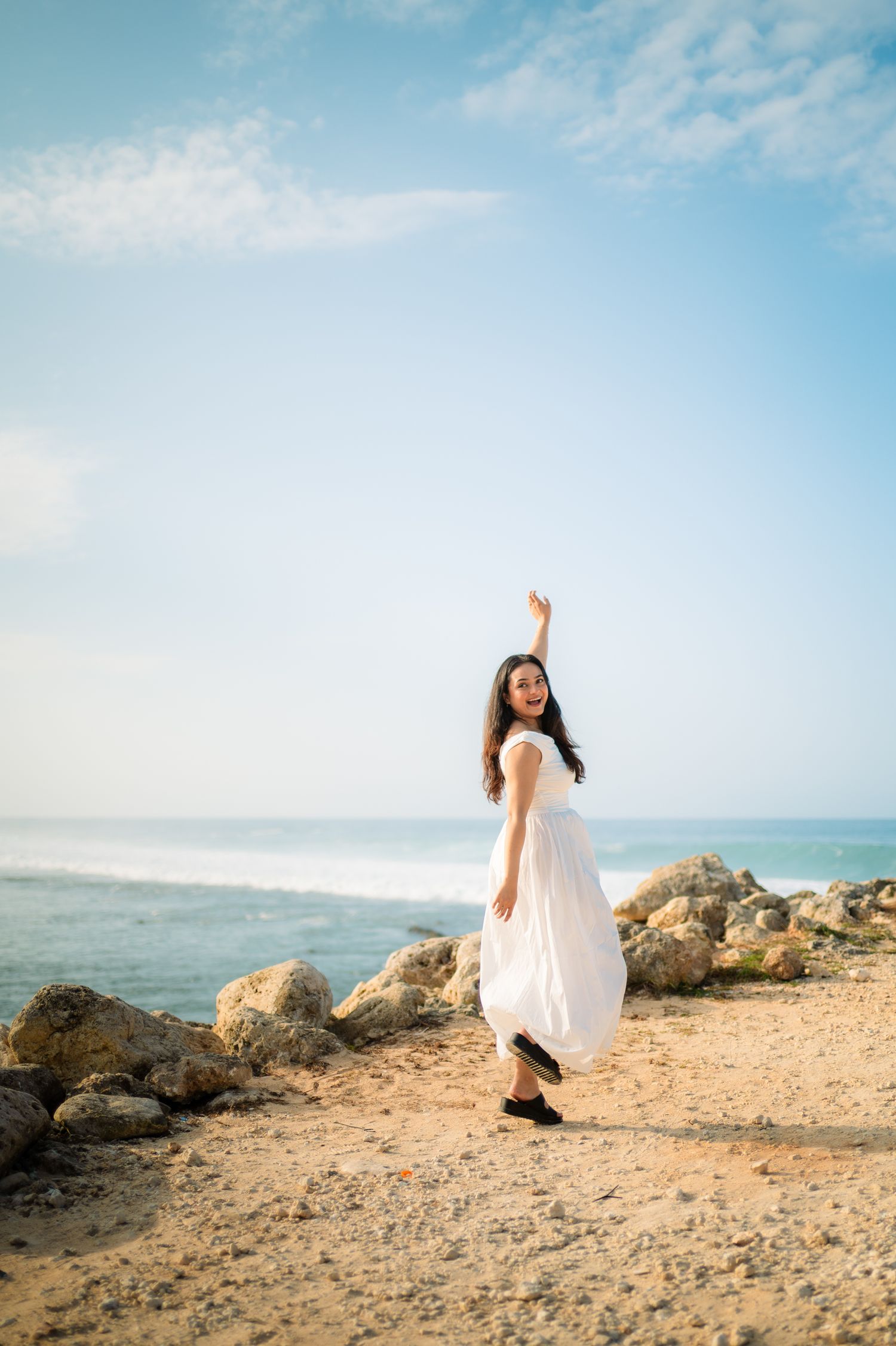 Woman in white dress posing on rocky beach with arm raised, ocean and blue sky in background.