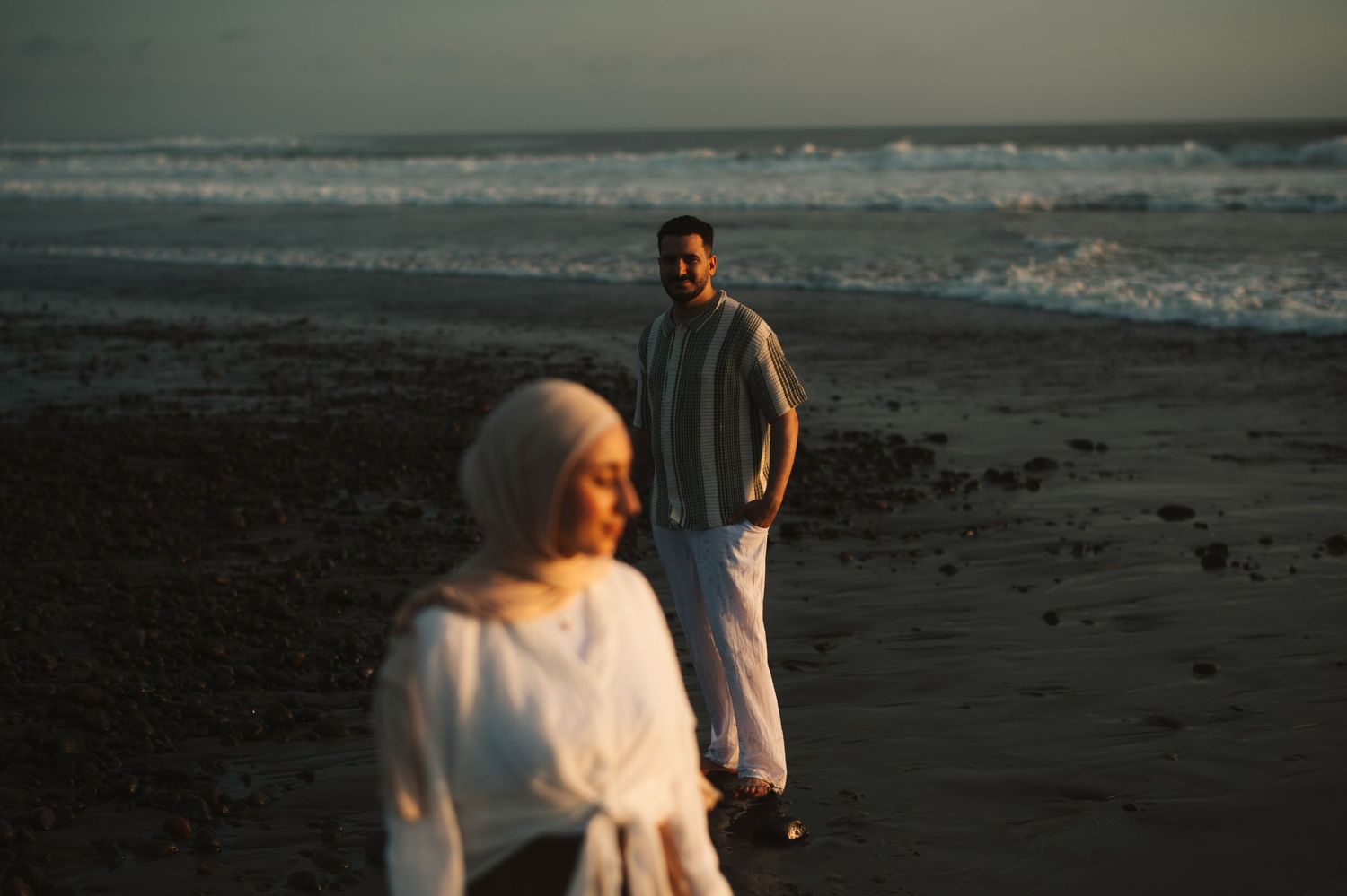 A couple in white attire stands on a sandy beach at sunset, with ocean waves in the background.