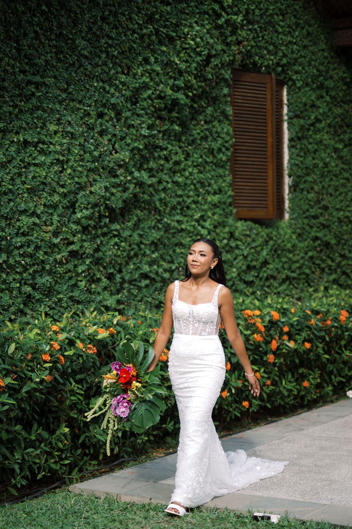 Elegant bride in fitted white lace gown walks past lush green ivy wall holding tropical bouquet.