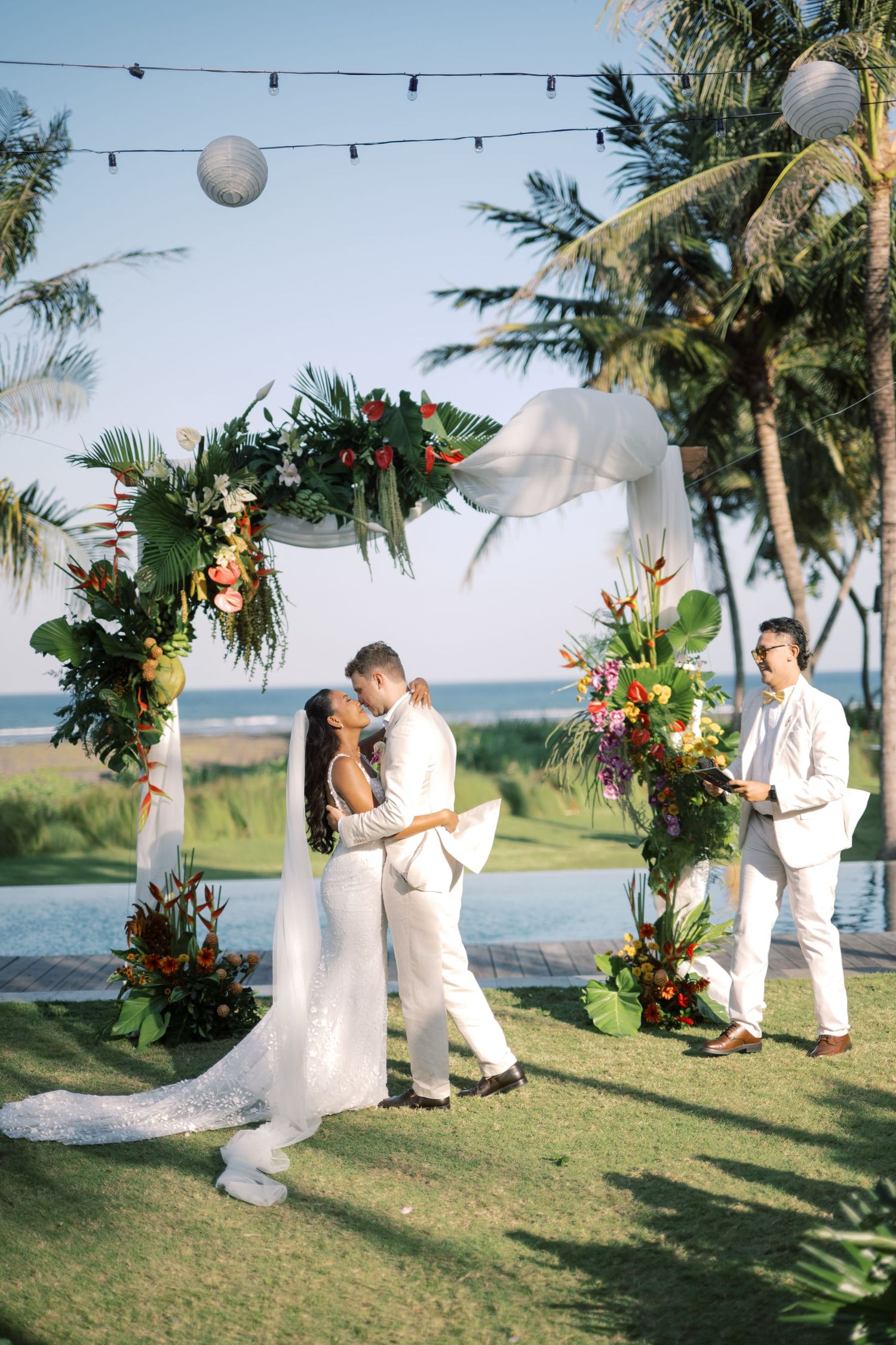 Bride and groom share first kiss under tropical floral arch at outdoor beach wedding ceremony.