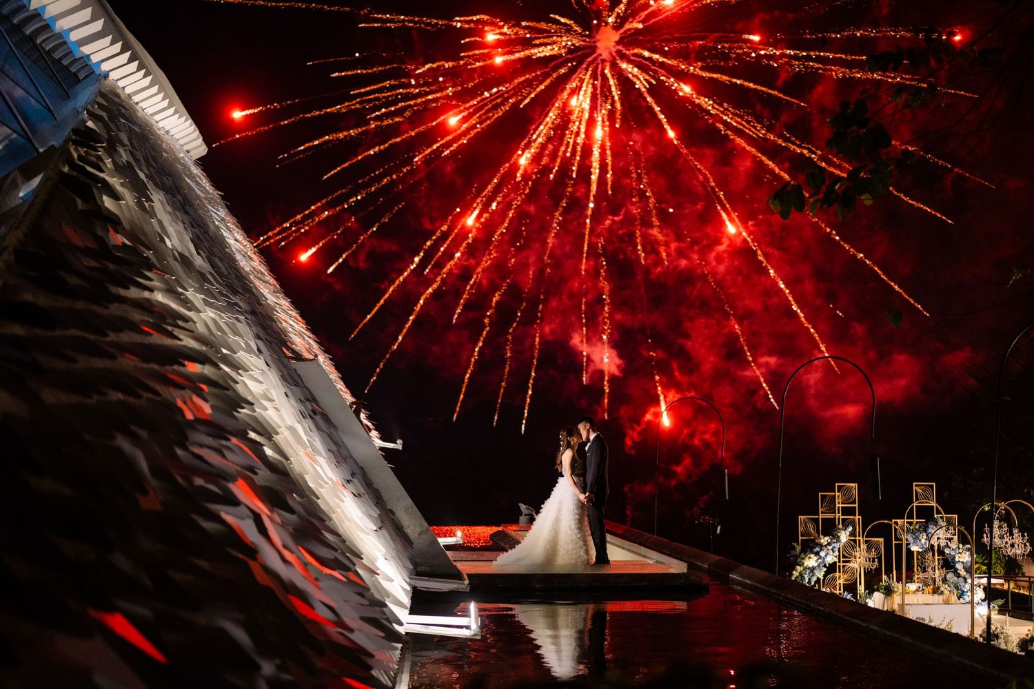 Bride and groom embrace on a waterfront platform at night as red fireworks burst across the dark sky above their wedding celebration.