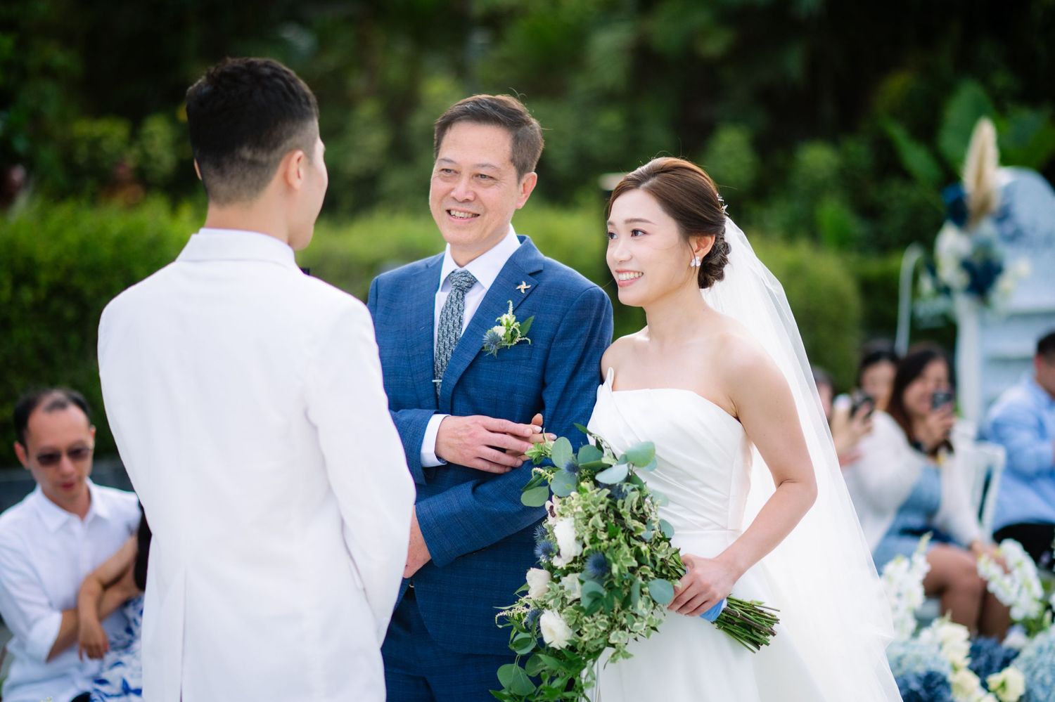 Smiling bride in a white gown holding a white floral bouquet stands before an officiant in a navy suit during an outdoor wedding ceremony.