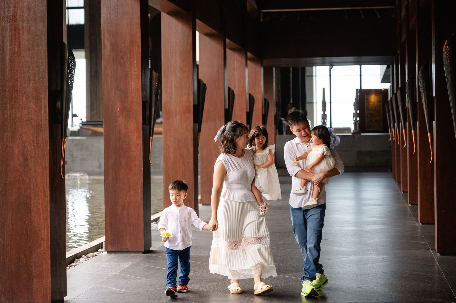 Family in white outfits walking through a wooden corridor with children, in a modern Asian architectural setting.