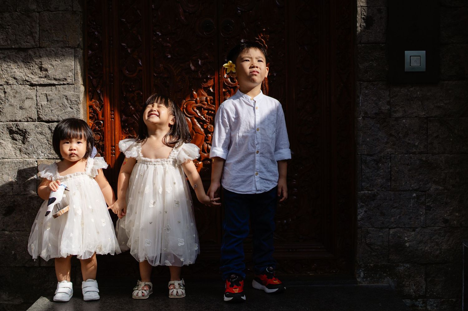 Three Asian children in formal attire pose together near an ornate wooden door in warm sunlight.