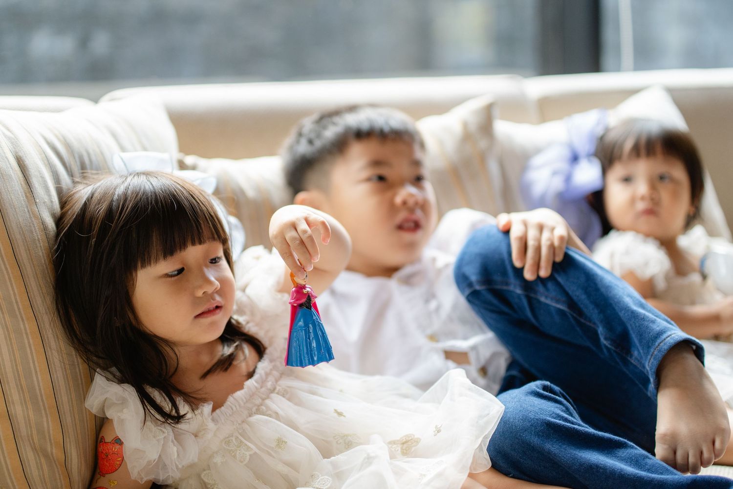 Three Asian children relaxing on a beige sofa, with a girl holding a blue tassel toy in the foreground.