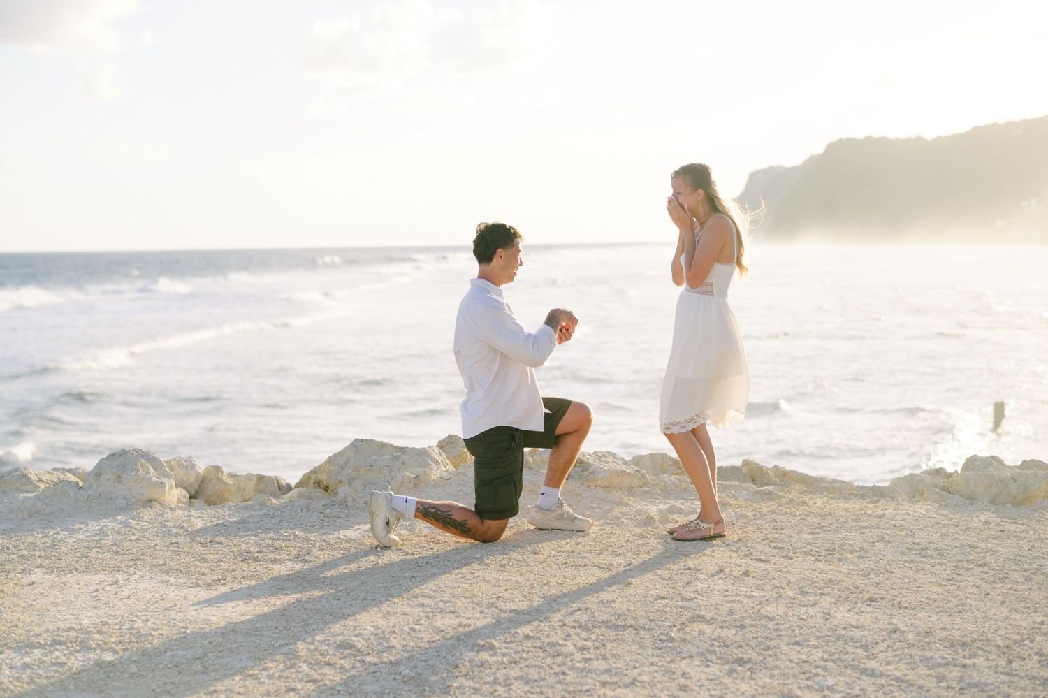 Man proposing to woman on rocky beach at sunset, holding ring box while kneeling, waves crashing in background.