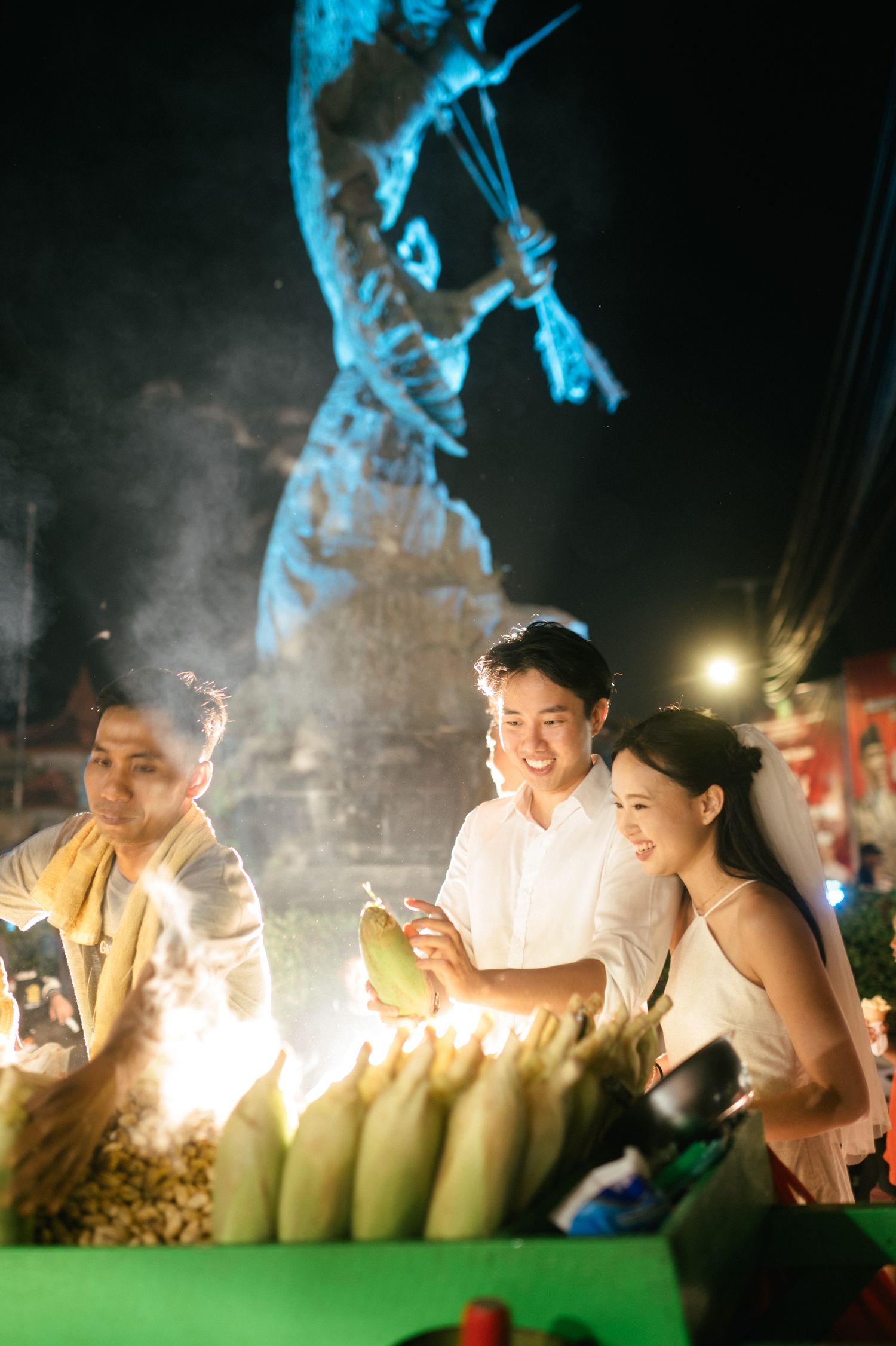 Couple smiling at a night market corn stall with glowing street lights and a lit statue in the background.