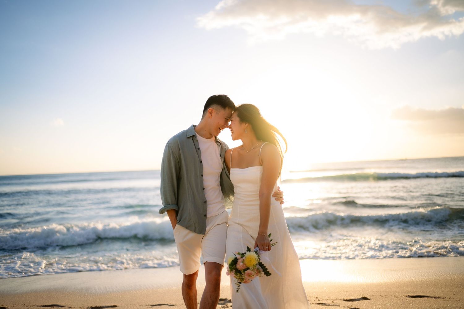 Bride and groom share a romantic moment on a sunny beach at sunset, waves crashing behind them.