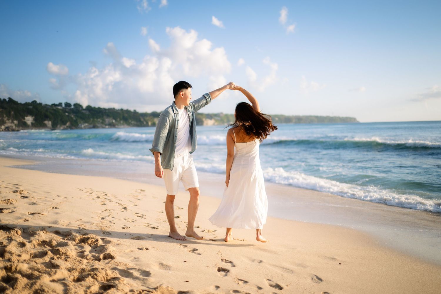 Couple dancing on a sunny beach with ocean waves, white sand, and lush green hills in the background.