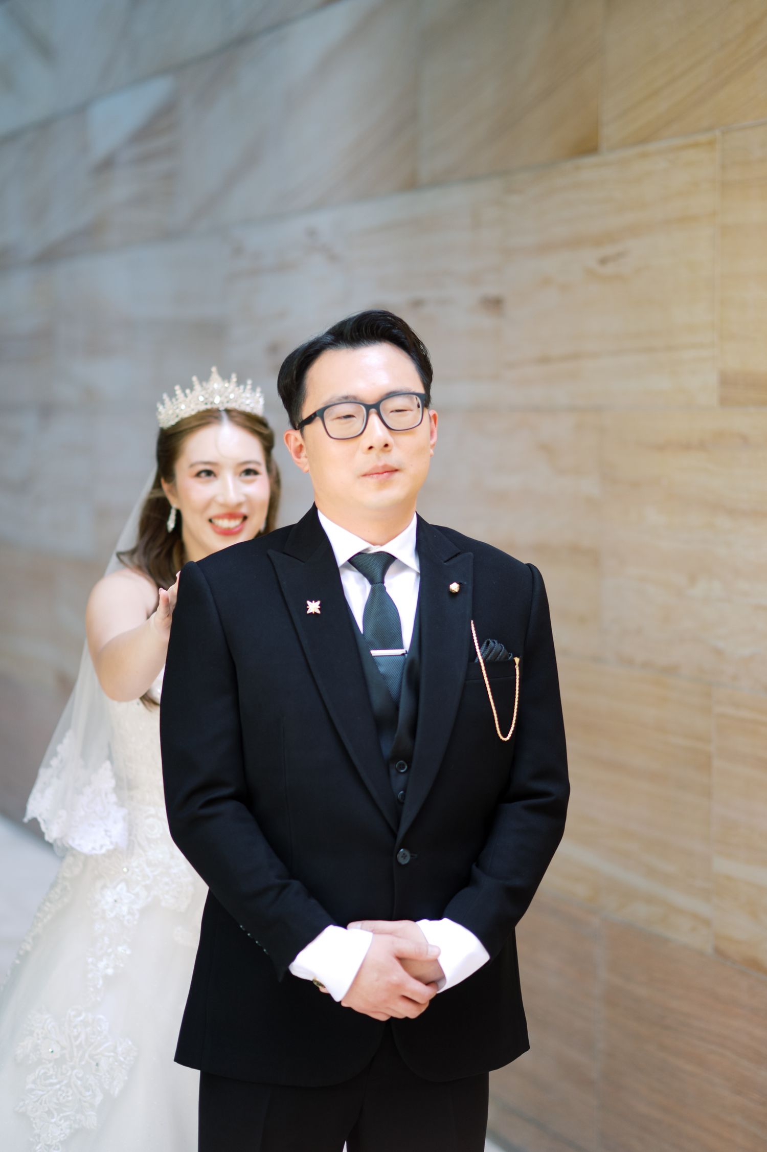 A bride in a white gown and tiara playfully peeks behind a groom in a black suit during their wedding photo session.
