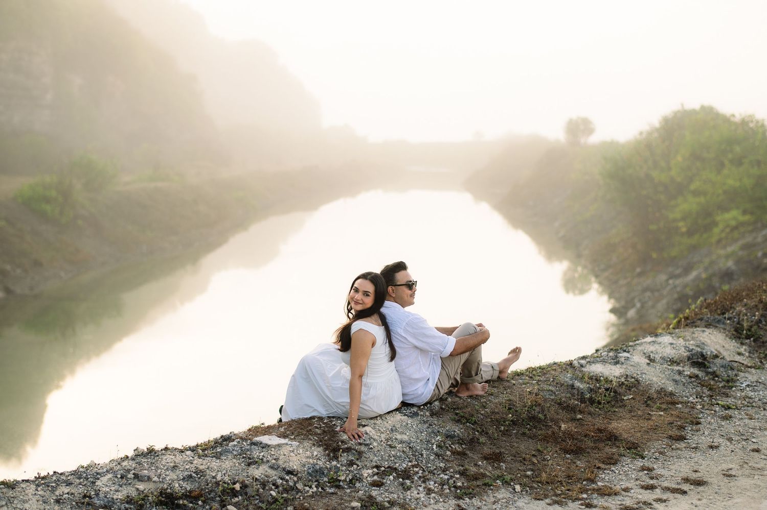 Couple in white sitting back-to-back on rocky cliff overlooking serene misty river in nature.