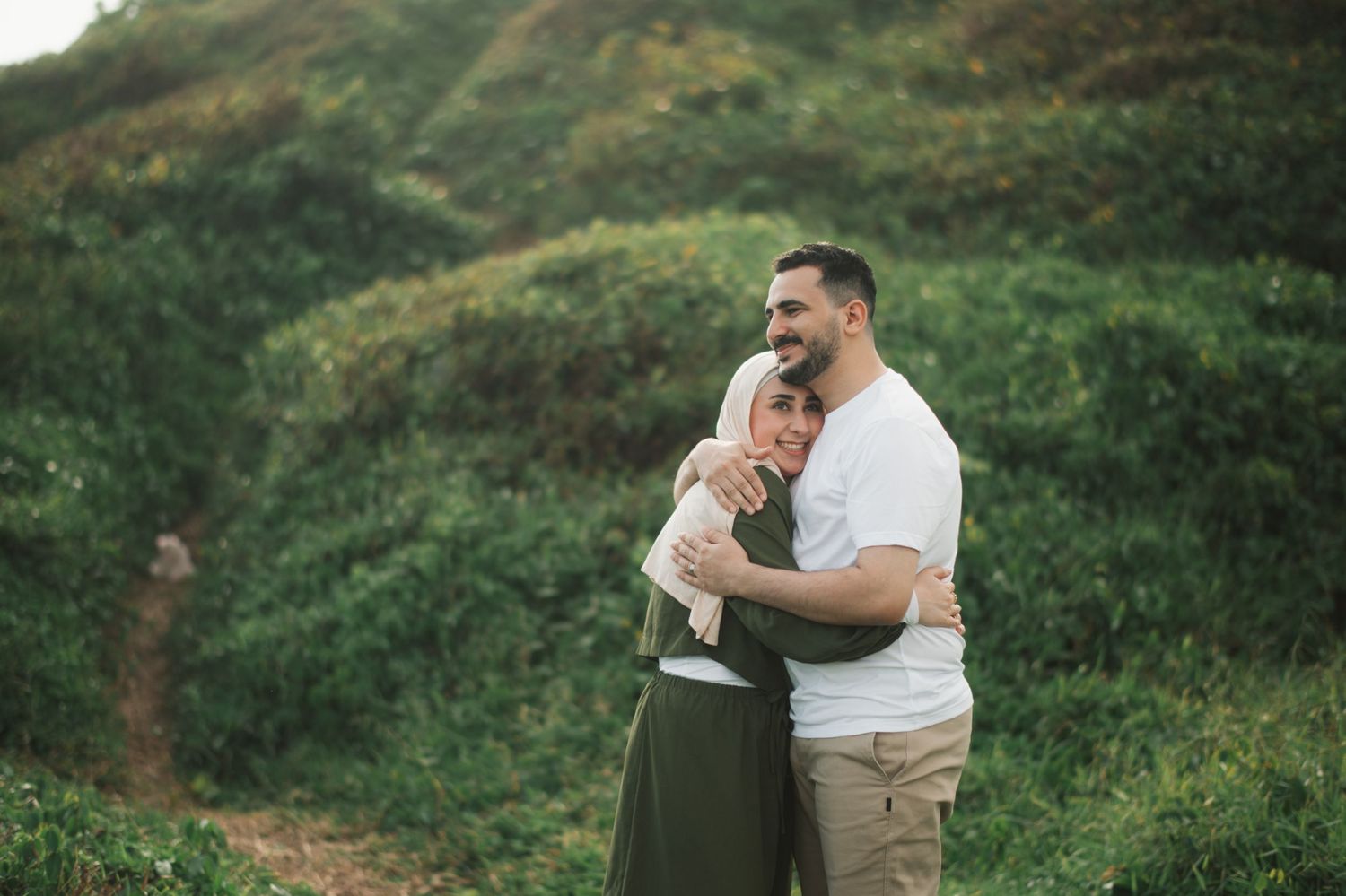 Couple embracing outdoors on a green hillside, man in white t-shirt hugging woman in hijab and olive dress.