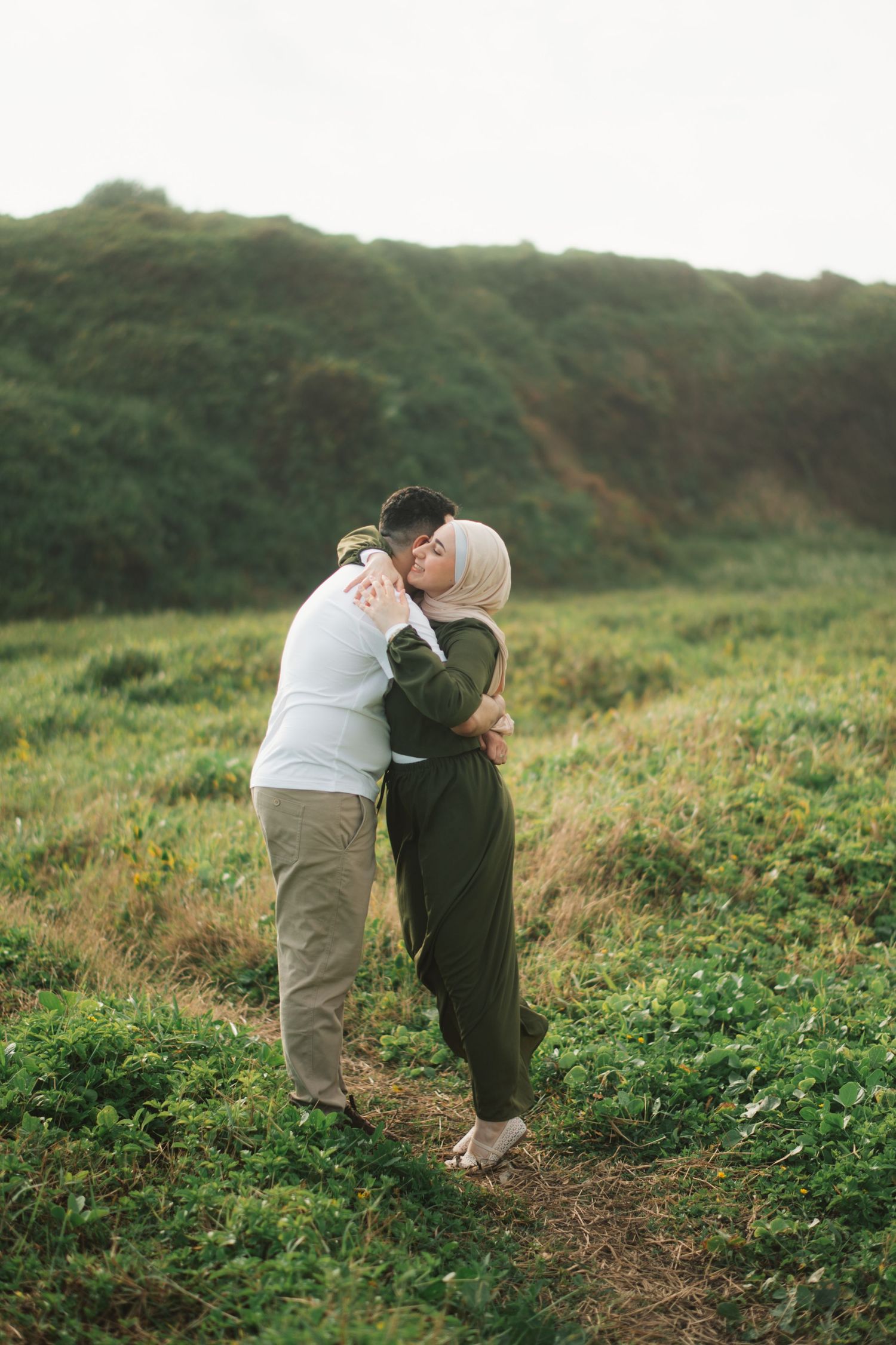 A couple embracing and kissing in a green meadow, woman wearing hijab and olive jumpsuit, romantic outdoor photo.