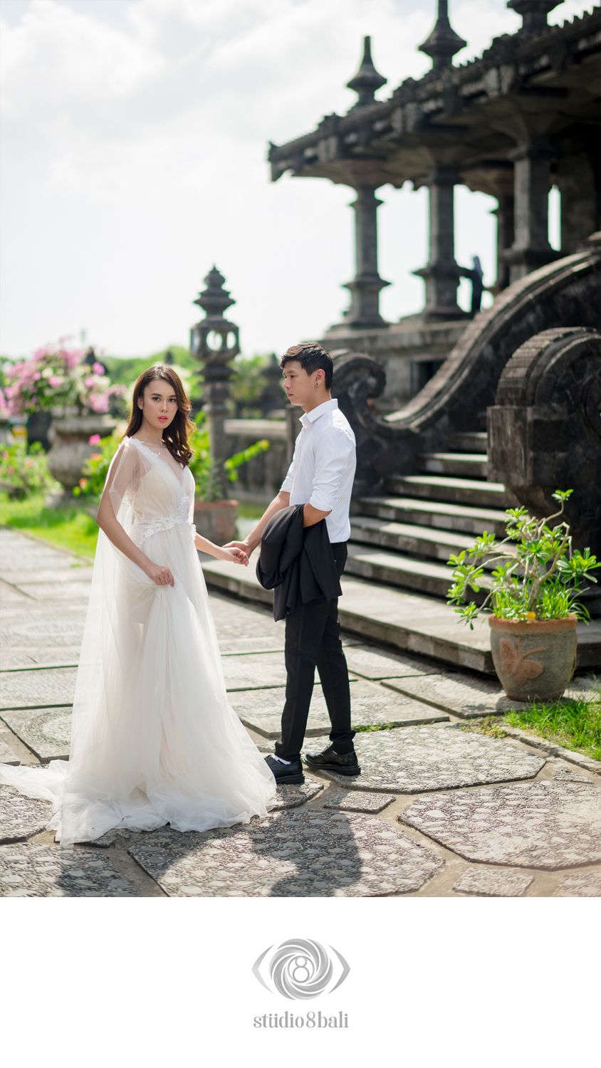 Couple in wedding attire holding hands at a Balinese temple, bride in white gown, groom in black suit, lush greenery.