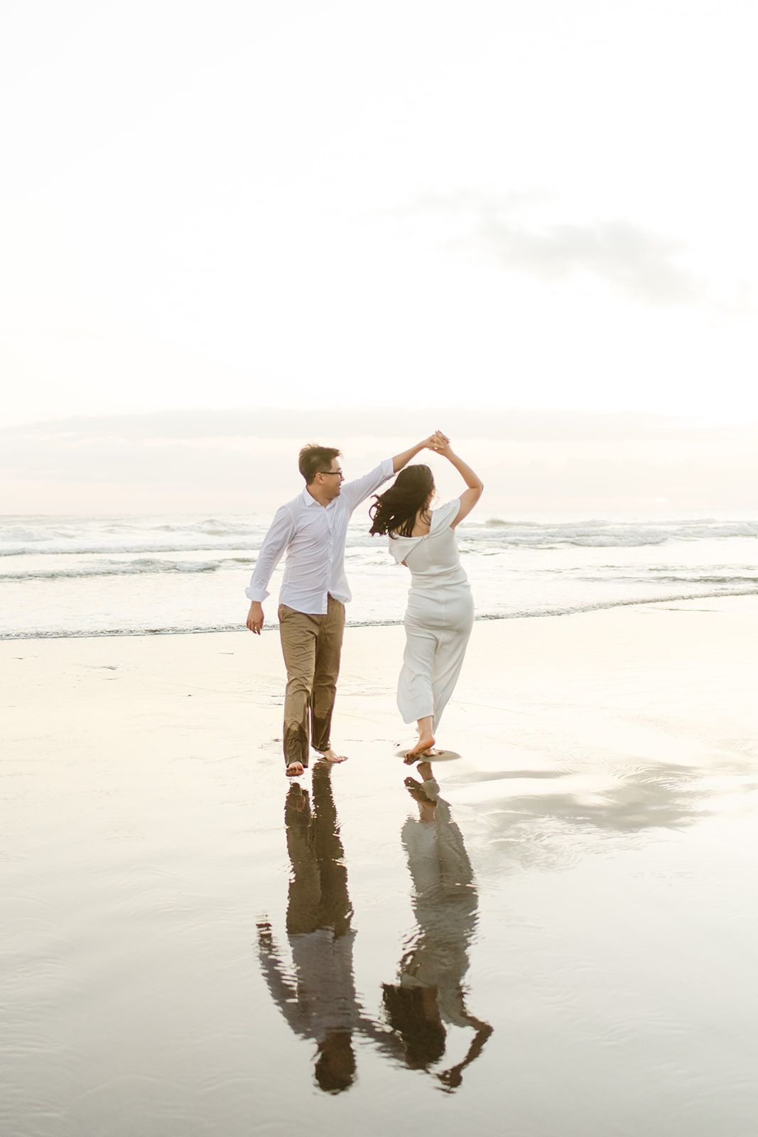 Romantic couple dancing on reflective wet beach at sunset, woman in white dress being twirled by man in white shirt.