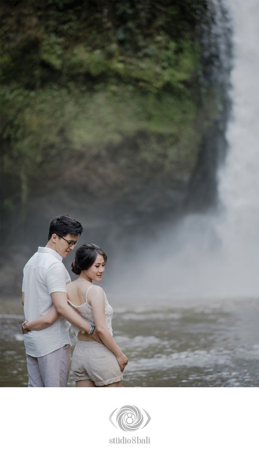 Romantic couple embracing in front of a misty waterfall, dressed in white, captured by Studio 8 Bali.