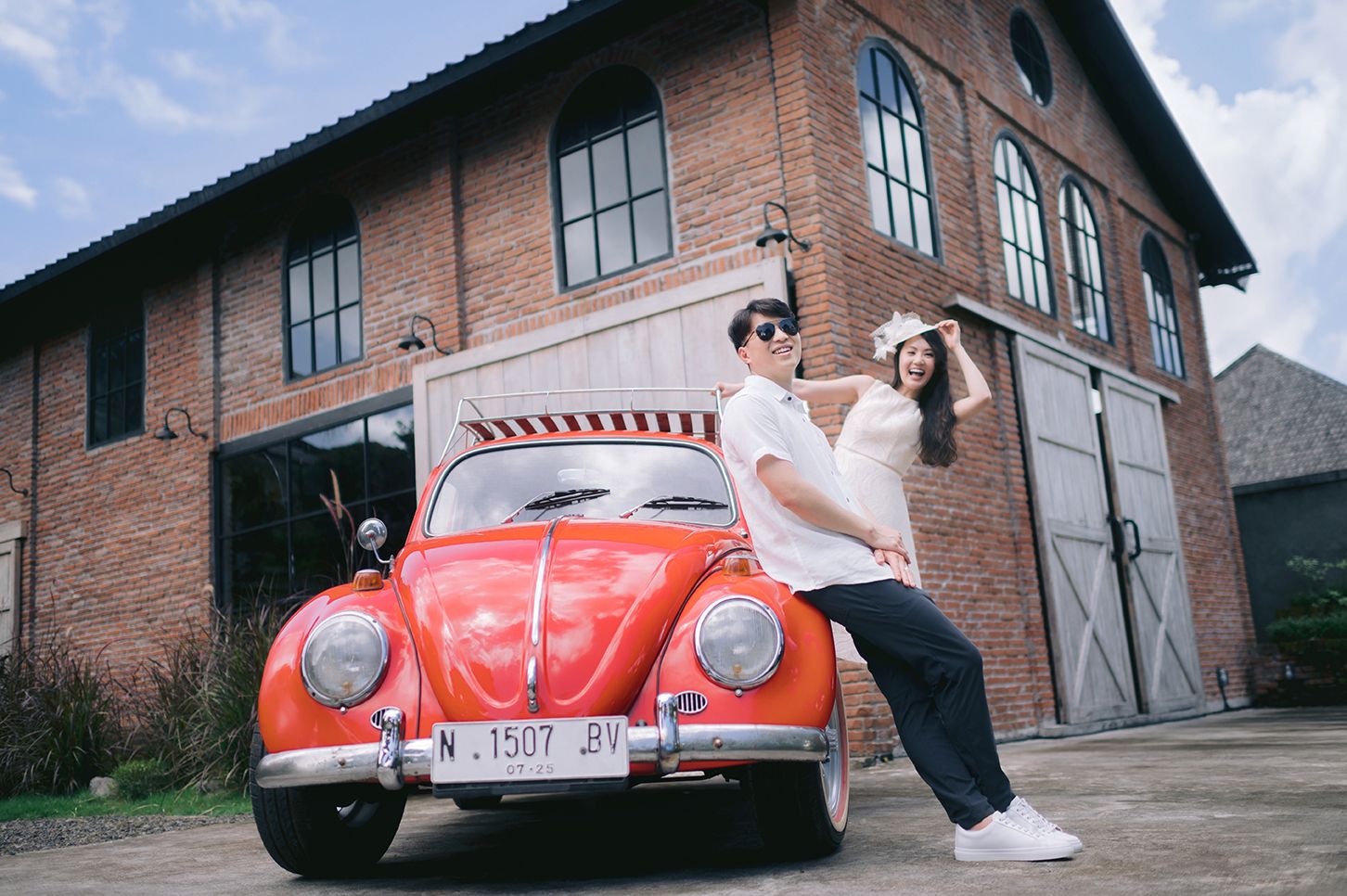 Couple posing with a vintage red Volkswagen Beetle outside a rustic brick building on a sunny day.