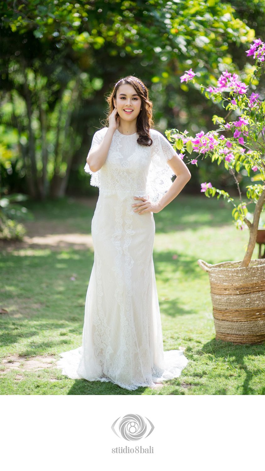 Bride in white lace wedding gown with cape sleeves posing outdoors in a garden with pink flowers.