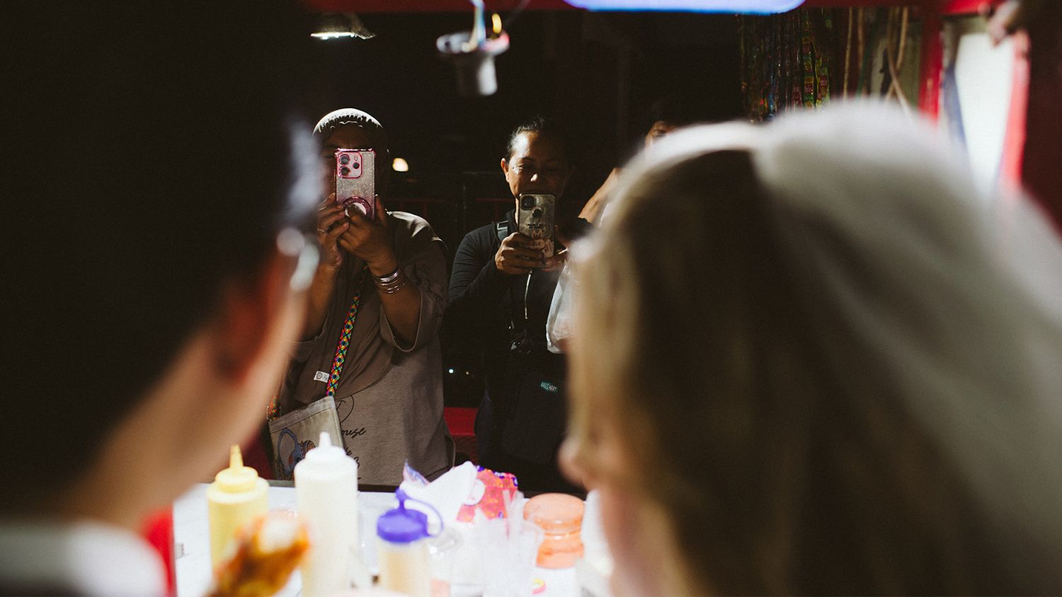 Man in suit photographs wedding guests at a reception table using a smartphone in a dimly lit venue.