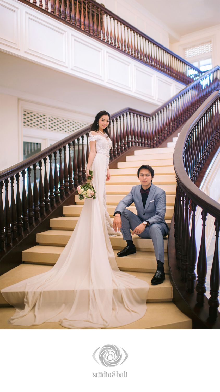 Elegant wedding couple posing on grand staircase, bride in white gown with train, groom in gray suit.
