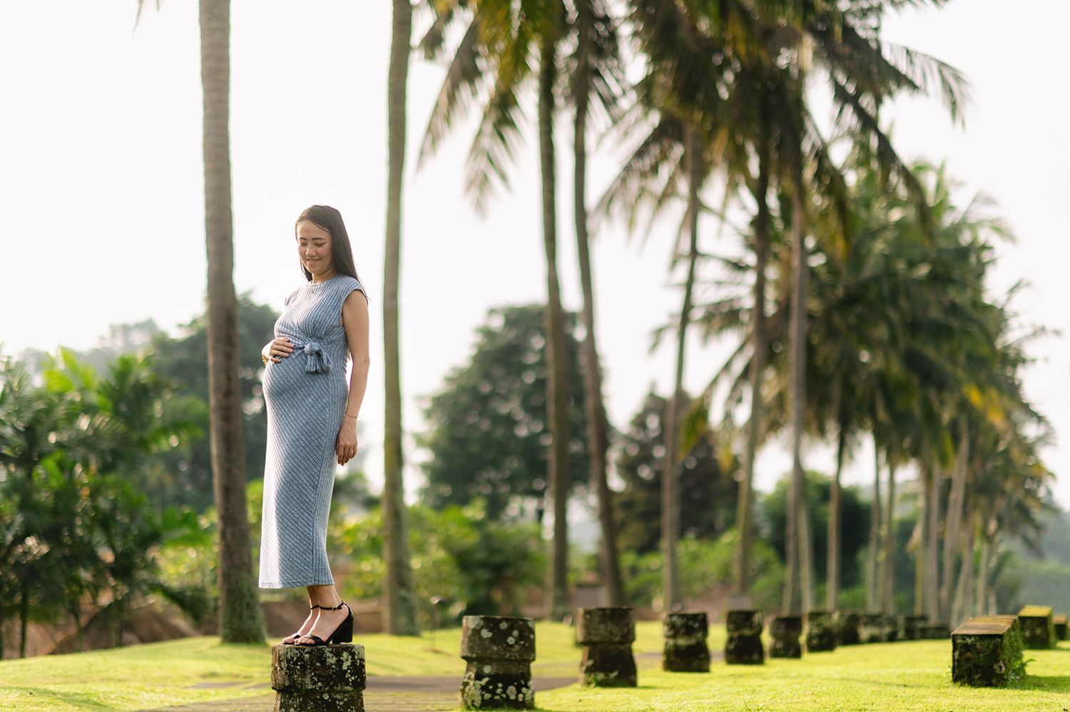 Pregnant woman in light blue dress standing on stone pillar surrounded by tall palm trees in tropical garden Maya Ubud Resort and Spa in Ba.