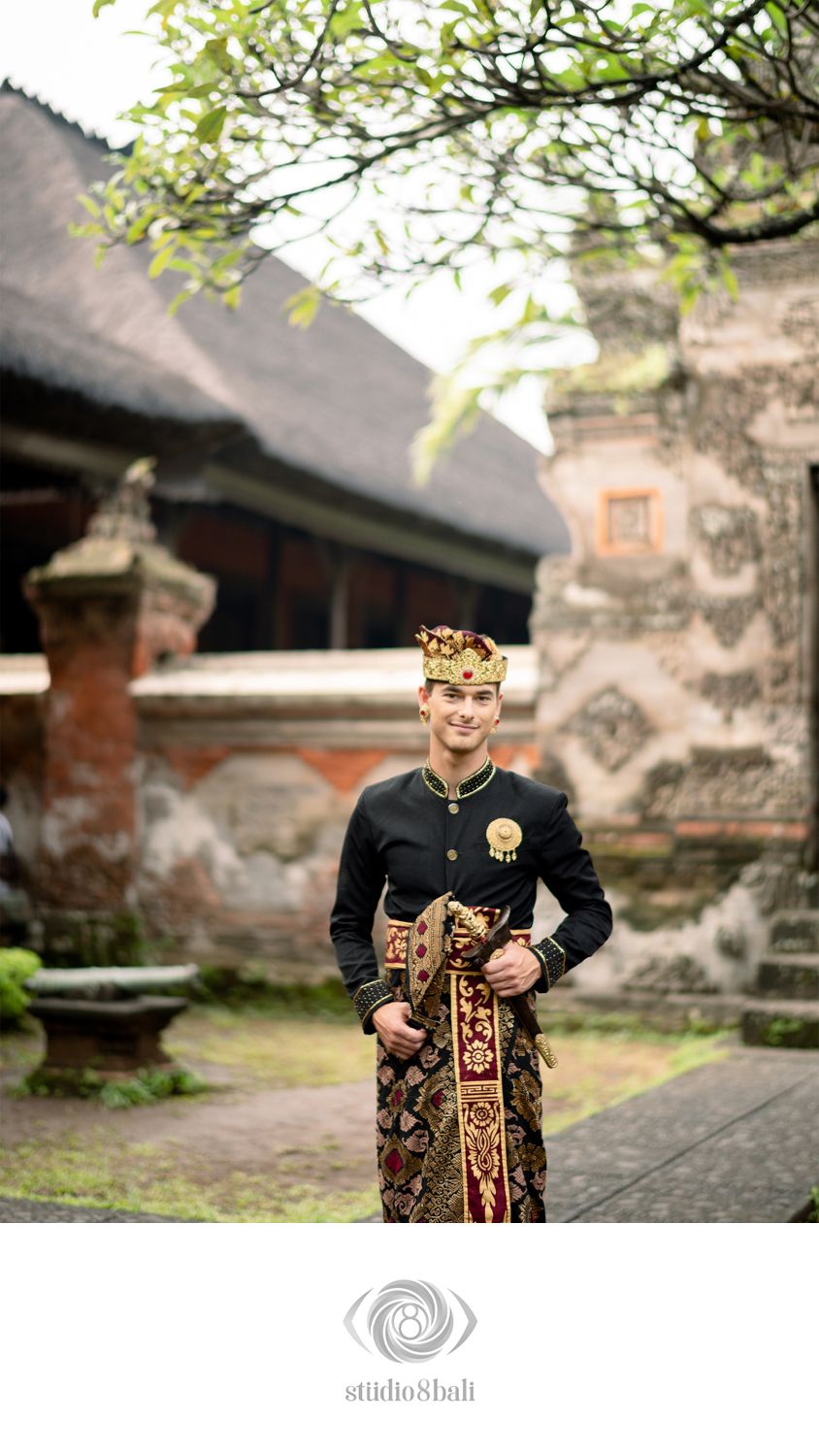 Balinese man in traditional black udeng crown and batik sarong stands proudly in a Balinese temple courtyard.