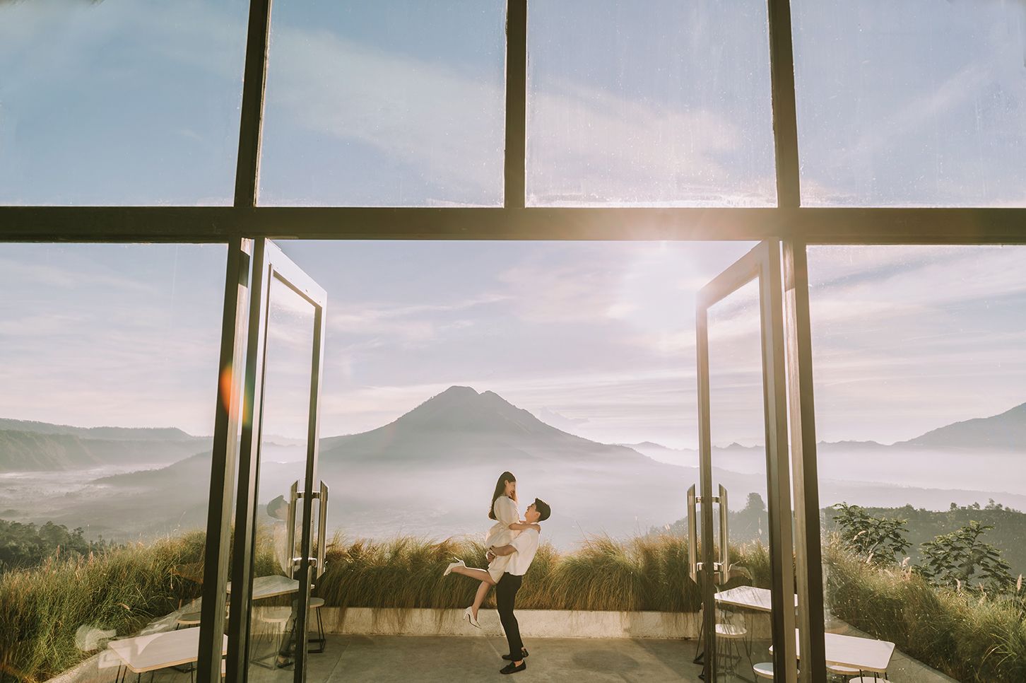 Couple embracing in a glass pavilion with stunning Mount Batur volcano and misty valley views in Bali.