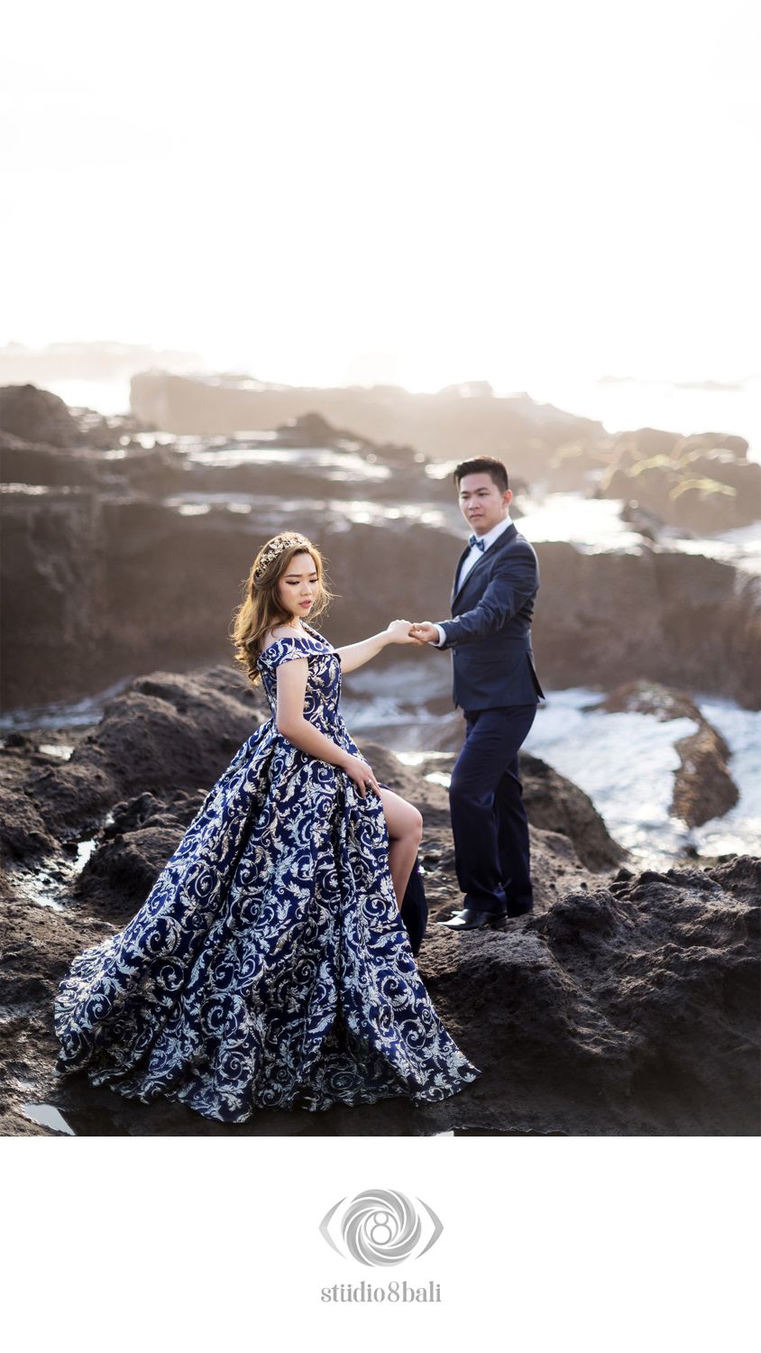 Couple in elegant attire pose on rocky coastal cliffs, woman in ornate black and white gown, man in navy suit.