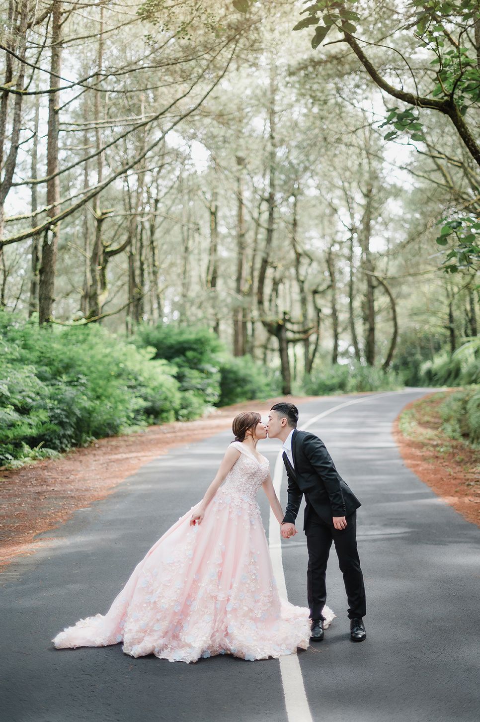 Couple sharing a romantic kiss on a tree-lined road, bride in pink floral gown and groom in black suit.