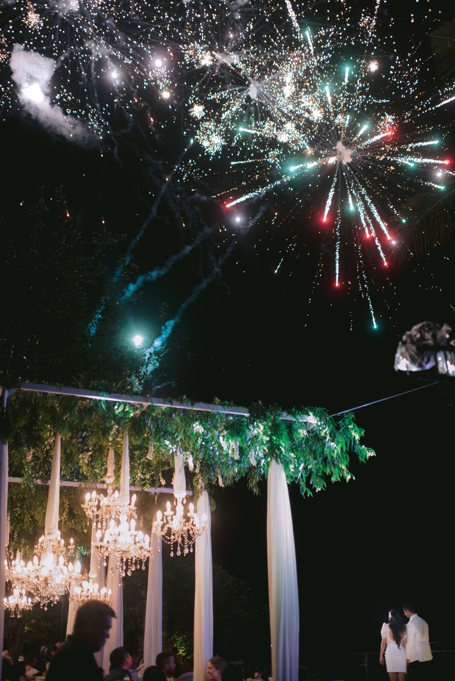 Colorful fireworks light up the night sky over an elegant outdoor wedding reception with chandeliers and floral garlands.