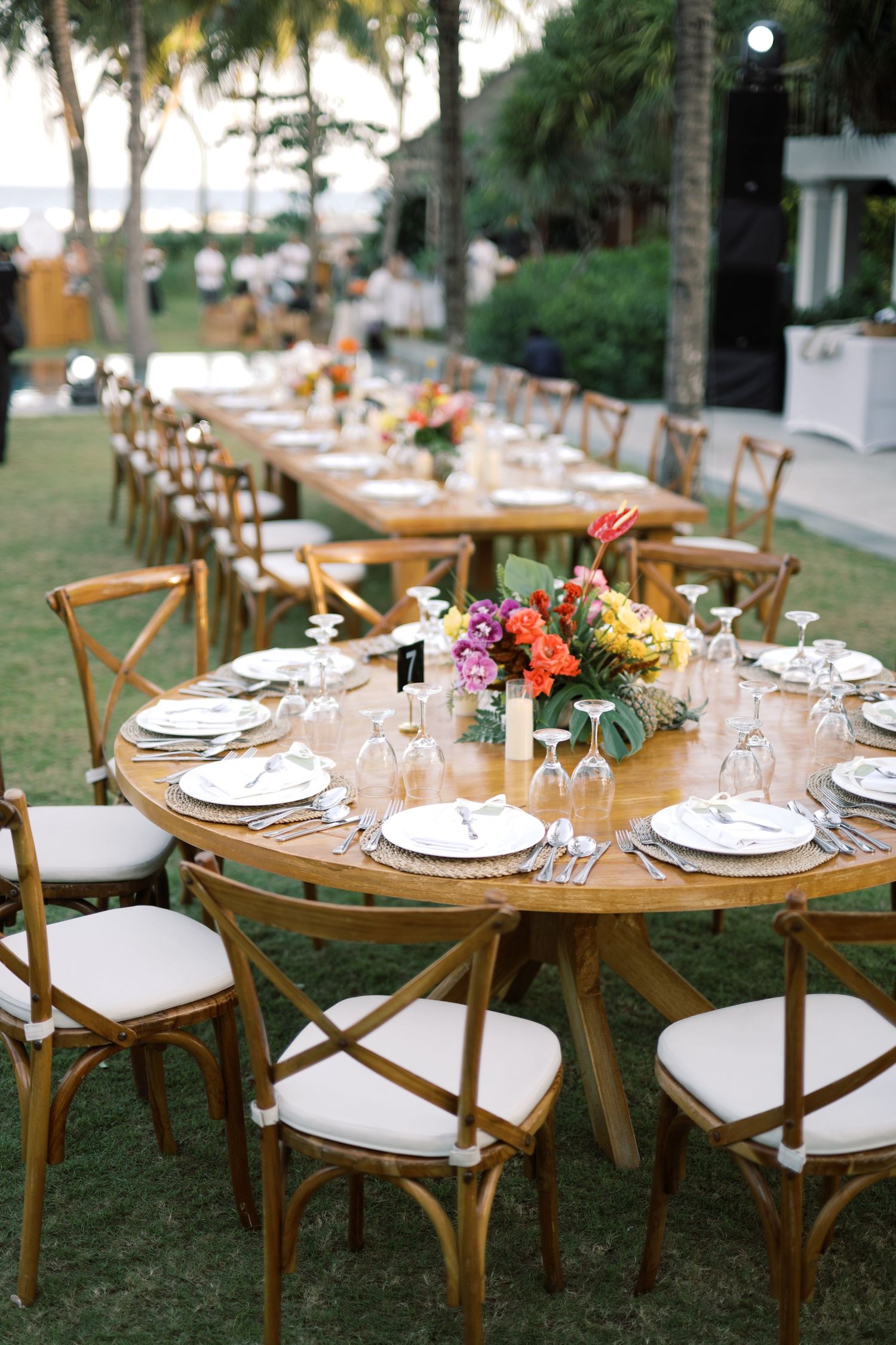 Elegant outdoor tropical wedding reception with wooden round tables, cross-back chairs, and vibrant floral centerpieces on lush green lawn.