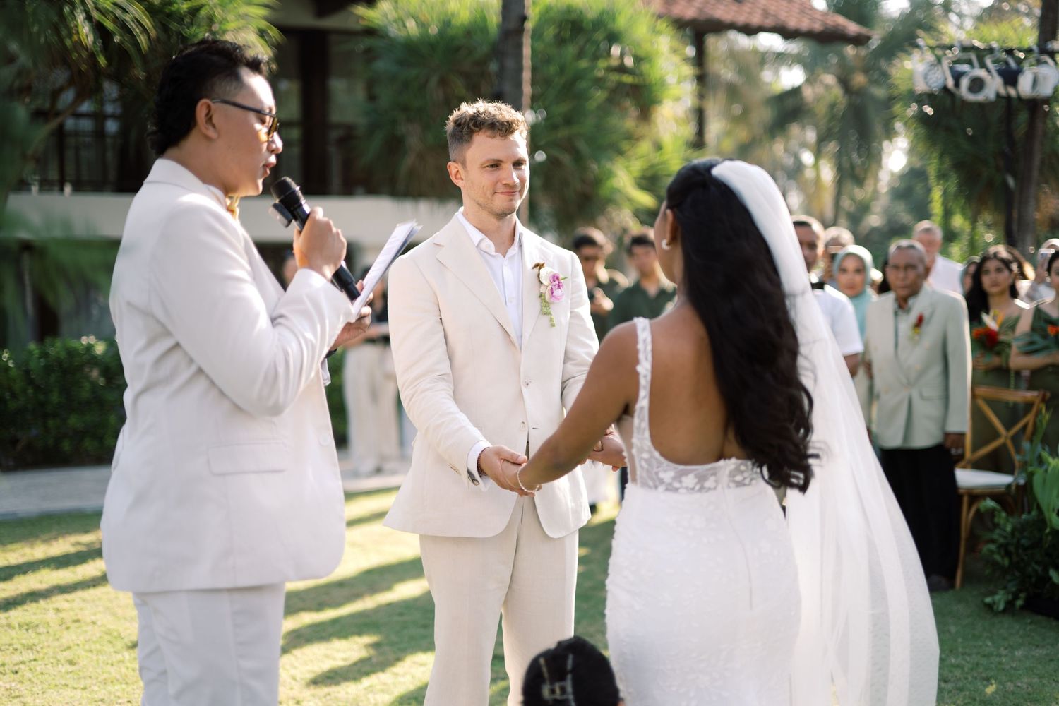 Groom in beige suit holds bride's hands during outdoor wedding ceremony with officiant speaking into microphone.