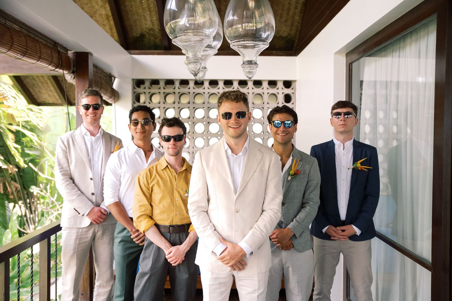 Groom in white suit poses with groomsmen in sunglasses at tropical wedding venue.