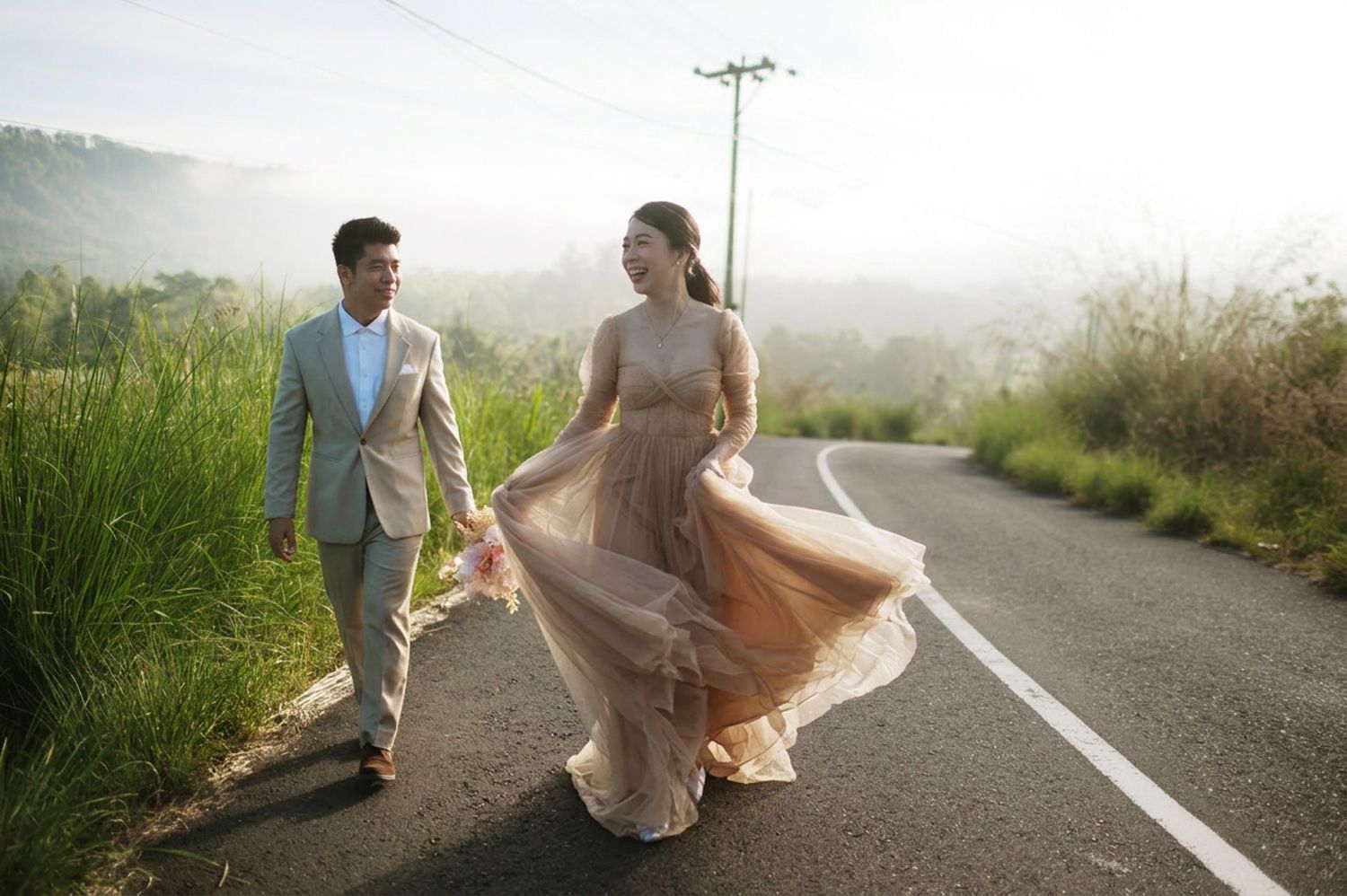 Couple in elegant attire walking hand-in-hand on a scenic road surrounded by lush greenery and misty mountains.