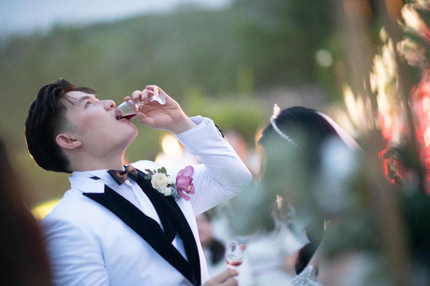 Groom in white tuxedo drinking champagne at outdoor wedding reception with floral boutonniere.