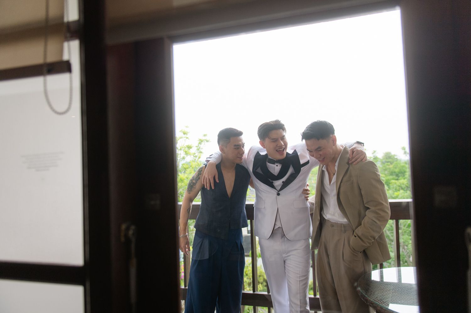Three men in formal attire share a warm embrace on a balcony surrounded by lush greenery.