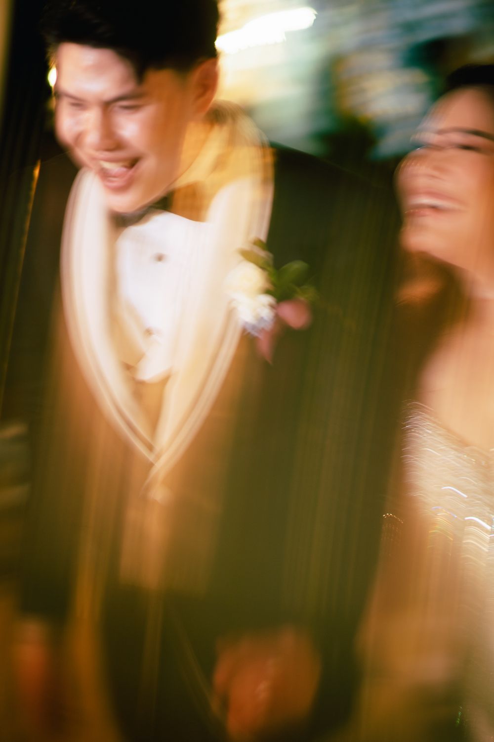 Joyful couple laughing together at wedding, groom in dark tuxedo with boutonniere, bride in elegant dress, motion blur effect.